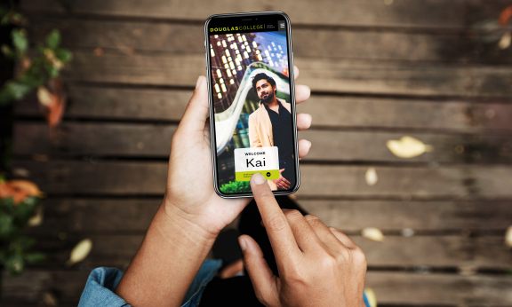 A person is holding a mobile phone with two hands. One finger is touching the screen of the phone. On the screen there is a photo of a young man and the words Welcome Kai.